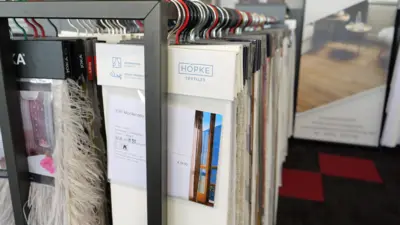 Nahaufnahme mehrerer hängender Stoff- und Materialmuster in einem Ausstellungsraum. Die Musterkarten zeigen unterschiedliche Texturen, Farben und Oberflächen und sind auf einer Stange angeordnet, mit weiteren Präsentationselementen im unscharfen Hintergrund.